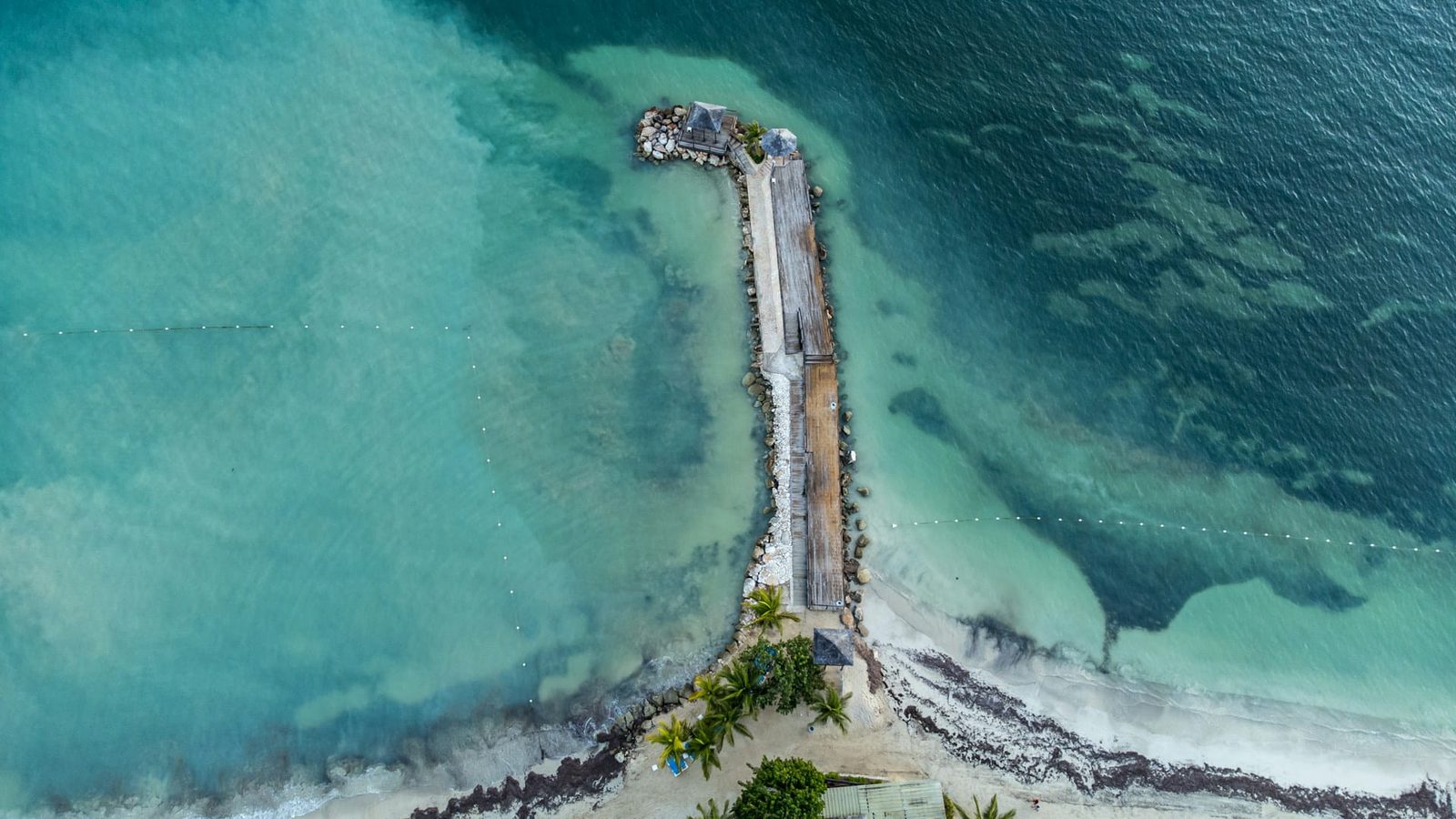 An aerial view of a wooden jetty reaching into turquoise water from a beach on Jamaica's north coast
