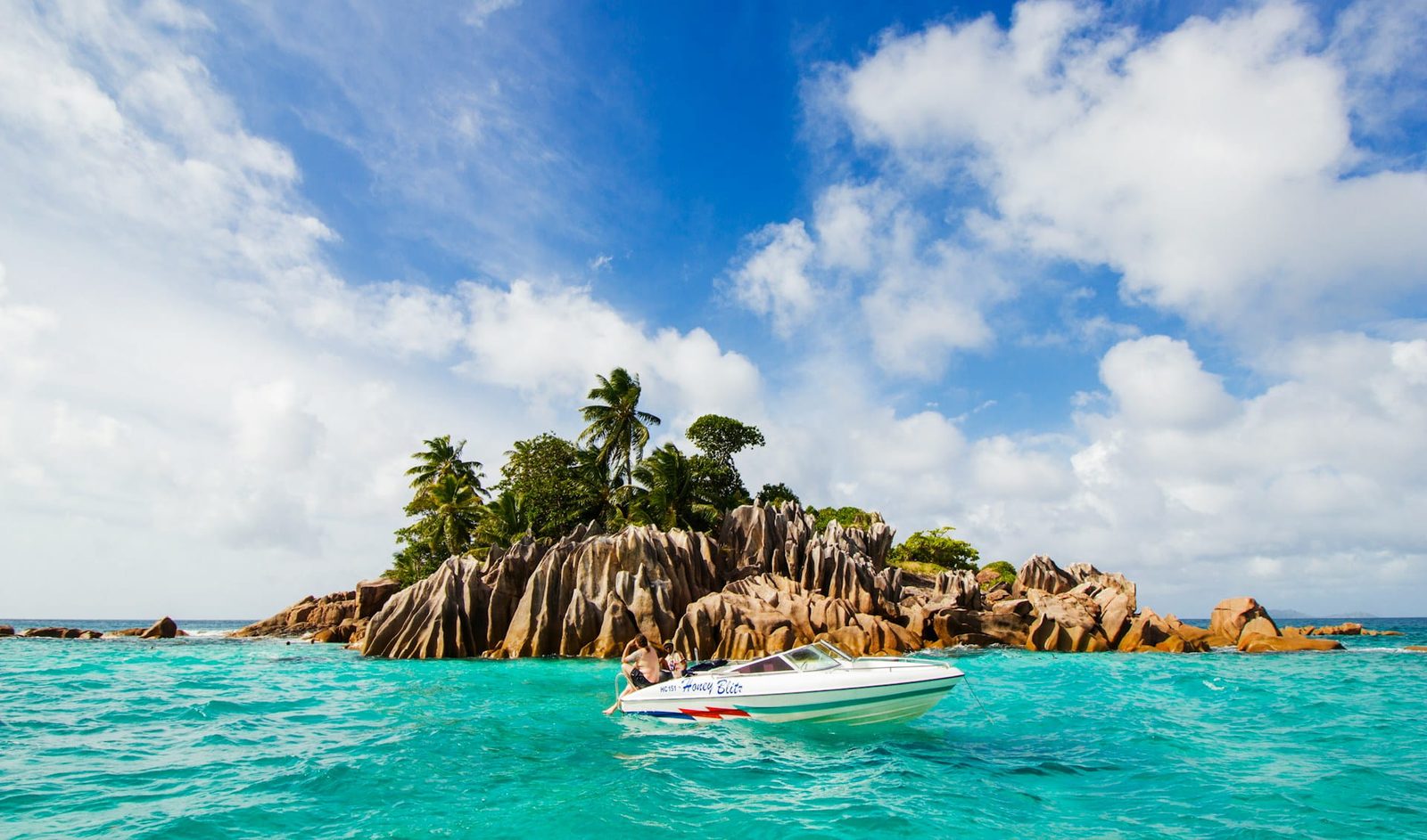 A speedboat in turquoise water beside a palm-topped granite islet in the Seychelles, showing the islands across the seasons