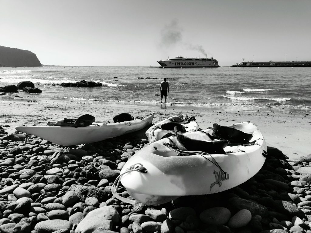 Monochrome beach view with kayaks, pebbles, and a ferry in the background.